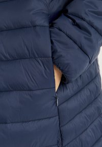 Marinblå pufferjacka med horisontell sömnad, en zip-fick på sidan och en slät, lätt tygkänsla.