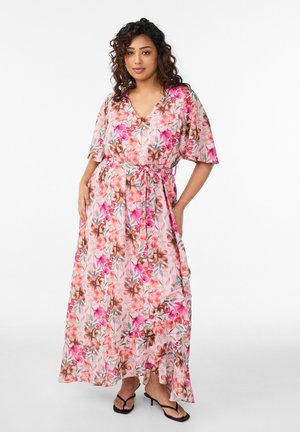 Femme portant une robe maxi fleurie rose à manches courtes et ceinture à la taille, debout les mains dans les poches, chaussée de sandales à talons noires.