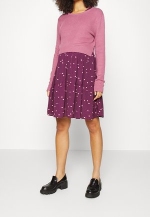 Kvinde iført en pink kort sweater, lilla nederdel med pink prikker og sorte chunky loafers, der står mod en hvid baggrund.