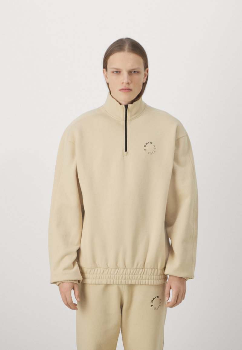 Felpa beige con zip al collo in tessuto morbido, con orlo a costine, cuciture laterali e un piccolo logo sul petto.