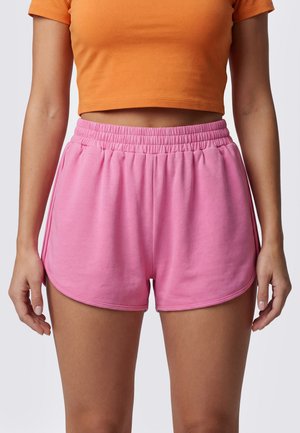 Vrouw die felroze shorts met elastische tailleband en een cropped oranje t-shirt draagt tegen een effen lichte achtergrond.