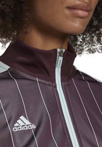 Bordeauxfarbene Trainingsjacke mit hellblauen Streifen und hohem Kragen. Verfügt über einen Reißverschluss vorne und ein gesticktes Adidas-Logo auf der linken Seite.