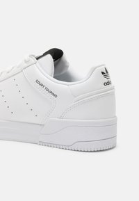 White leather sneaker with perforated details, black heel tab, padded collar, and textured rubber sole. Features printed branding on the side.