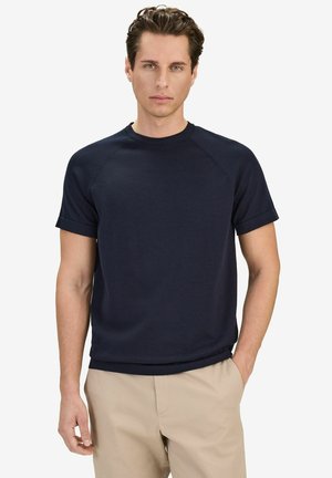 Man met kort krullend haar, gekleed in een marineblauw shirt met korte mouwen en beige broek, staand met één hand in de zak.