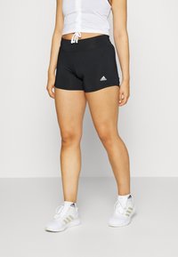 Pantalones cortos deportivos negros con una textura suave, que presentan una cintura alta y un logo sutil en el dobladillo izquierdo. Llevados con zapatillas blancas.