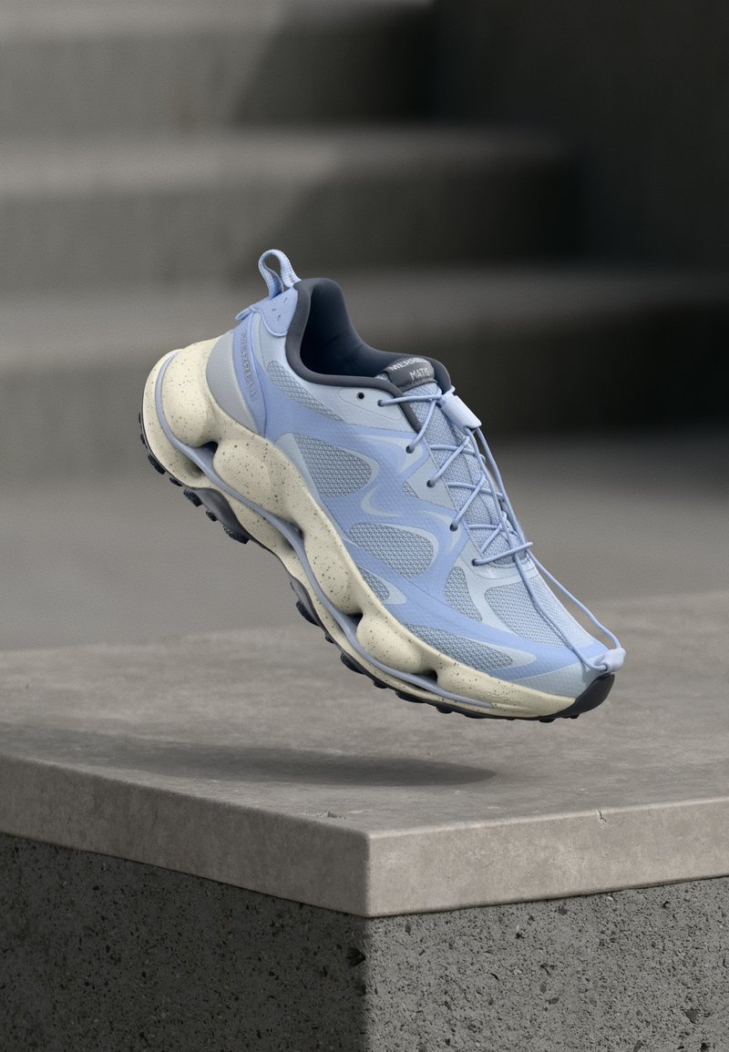 Αθλητικό παπούτσι σε ανοιχτό γαλάζιο με σχεδίαση που μοιάζει με κάλτσα και σόλα σε μπεζ με στίγματα, τοποθετημένο στον αέρα πάνω από επιφάνεια από μπετόν και σκαλοπάτια στο φόντο.