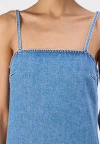 Vaaleansininen farkkutoppi ohutolkaimilla, jossa on neliön muotoinen pääntie ja minimalistinen muotoilu. Eroava neulontakuvio näkyy reunoilla.