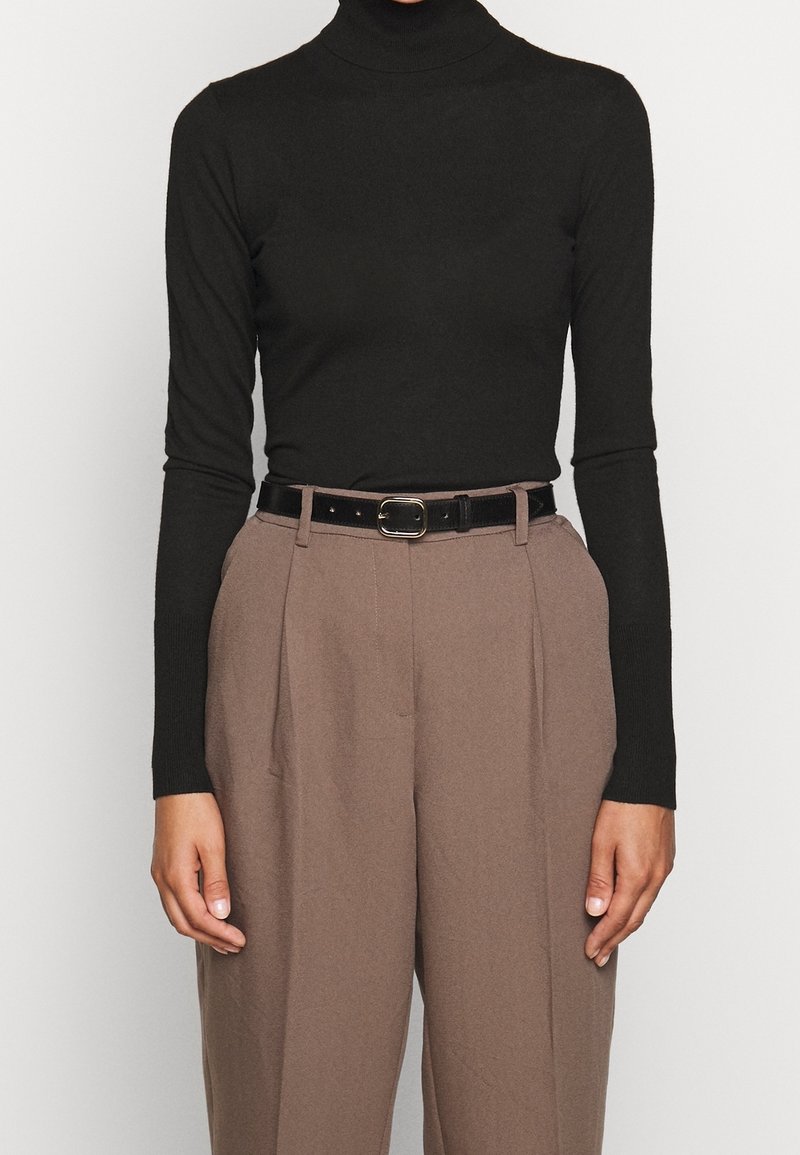 Frau in einem schwarzen Rollkragenpullover, der in braune hochgeschnittene Hosen mit Falten und einem schwarzen Gürtel mit goldener Schnalle gesteckt ist.