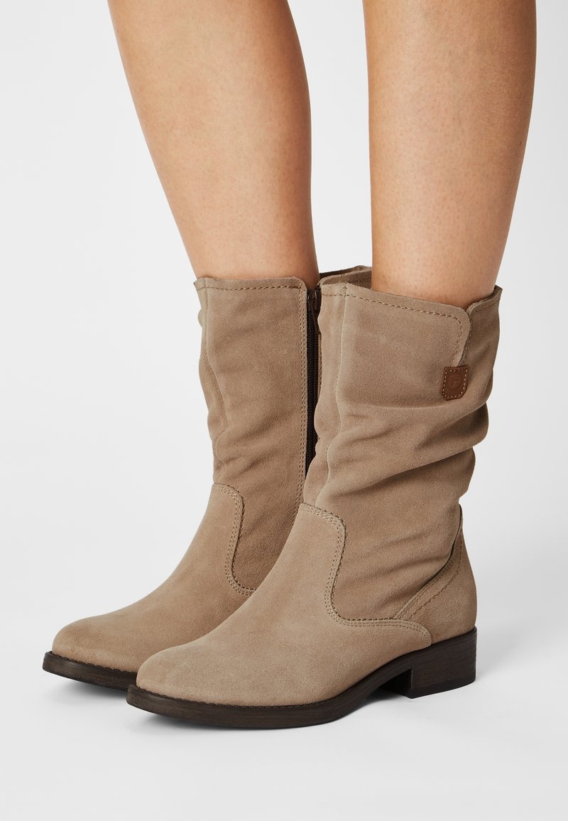 Bottes en daim beige mi-mollet avec fermeture éclair latérale et petit talon empilé, portées sur des jambes nues, design légèrement bouffant du shaft.