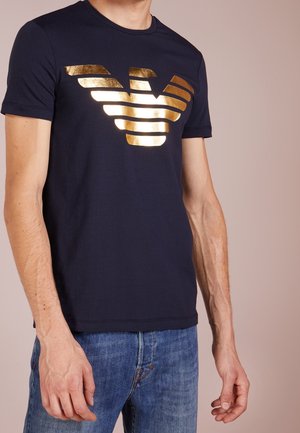 Navy blue t-shirt made of cotton with a crew neck. Features a metallic gold logo design at the front in striped patterns.