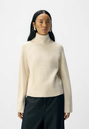 Femme aux longs cheveux noirs portant de grandes boucles d'oreilles blanches, un pull à col roulé crême à côtes et un pantalon rayé sombre, se tenant contre un fond uni.
