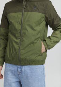 Olive green lightweight jacket with a zip front, elastic cuffs, and waistband. Features a contrasting darker green panel at the shoulders.