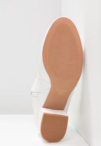 Botín blanco angular contra una pared, mostrando su suela texturada marrón y tacón cuadrado, talla 37, con la marca "ANNA FIELD."