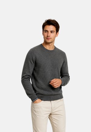 Young man wearing a dark gray long-sleeve sweater and light beige pants, with one hand in his pocket and looking to the side.
