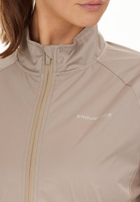 Ljust beige jacka med hög krage, framdragkedja och slät yta. Har "ENDURANCE"-logotypen i vitt på vänster sida.