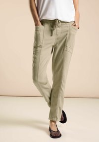 Pantalones de mezcla de algodón ligeros y beige con cinturilla elástica, bolsillos laterales y detalles de costura en contraste. Combinados con zapatos de malla negros.