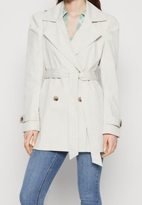 Trench coat beige claro con botones dorados de doble botonadura, solapas anchas y cintura ceñida con cinturón. Presenta mangas abullonadas y una textura suave.