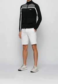 Svart zip-up jacka med texturerade detaljer, vita randiga accenter, tillsammans med vita shorts och ljusgrå sneakers.