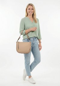 Frau trägt hellgrüne Bluse, hellblaue Jeans, weiße Turnschuhe und hält eine beige Schultertasche, steht auf weißem Hintergrund und lächelt.