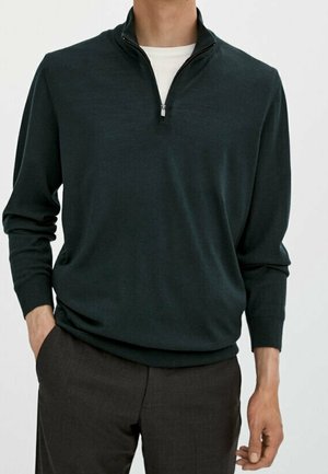 Homme portant un pull vert foncé à manches longues avec fermeture éclair jusqu'à la poitrine sur une chemise blanche, pantalon marron foncé, une main dans la poche, fond blanc.
