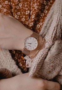 Reloj de malla en oro rosa con una esfera blanca minimalista, acompañado de un lazo de tela con textura en marrón claro con lunares y una pulsera.