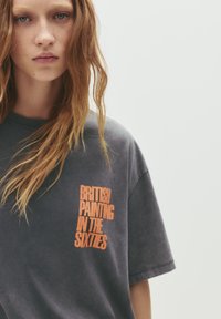 Mørkegrå bomulds T-shirt med korte ærmer. Har en orange grafik tekst, der står "BRITISH PAINTING IN THE SIXTIES" på forsiden.