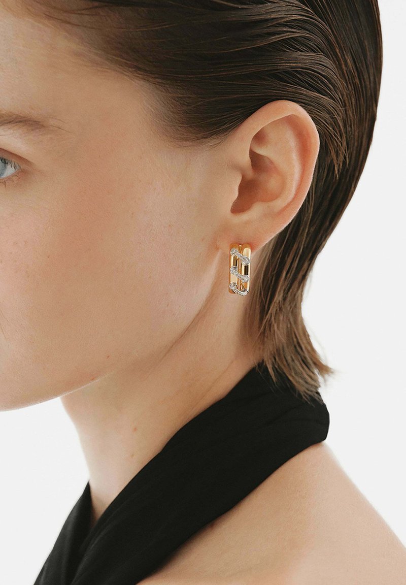 Pendientes en tonos dorado y plateado con un diseño de cadena entrelazada y pequeños acentos de diamante, llevados en la oreja sobre una tela oscura.