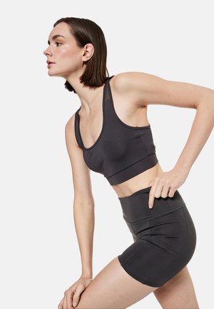 Jonge vrouw leunt naar voren, draagt een zwarte sportbeha en zwarte atletische shorts met hoge taille tegen een witte achtergrond.