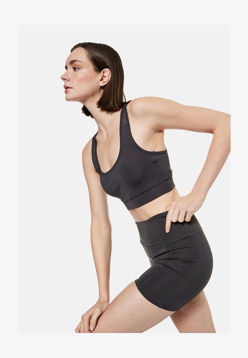 Jeune femme penchée vers l'avant, portant un soutien-gorge de sport noir et un short de sport noir taille haute sur fond blanc.