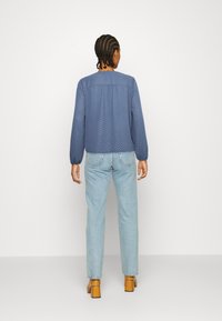 Blusa azul com bolinhas brancas e mangas elásticas, combinada com jeans azul claro e sapatos de salto bloco marrons. Fundo branco liso.