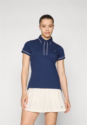 Navy blue polo shirt with white trim, five-button placket, and logo on the chest; worn with a pleated cream skirt. Smooth fabric texture.