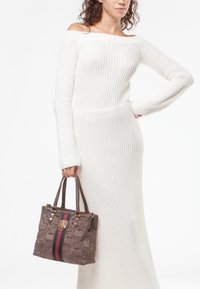 Bolso marrón con estampado y detalles en dorado, y una franja roja. Tela texturizada y asas resistentes. Combinado con un vestido blanco de tirantes al hombro.