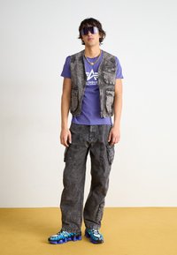 Gray denim vest over a purple T-shirt with a graphic. Worn with loose gray cargo pants and blue and black patterned sneakers.
