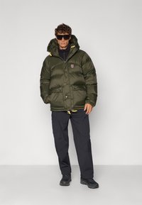 Doudoune vert foncé avec capuche, design matelassé, poches latérales, cordons de serrage jaunes et patch logo. Portée avec un pantalon noir et des chaussures.