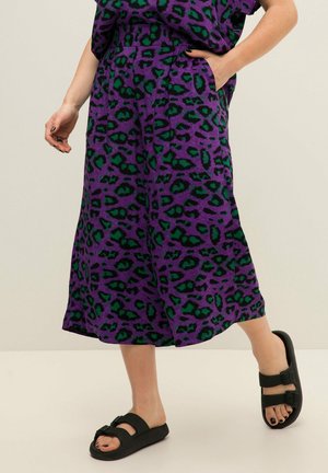 Jupe midi imprimé léopard violet et vert portée avec des sandales noires à bout ouvert et à boucle, une main dans la poche, debout sur un sol clair.