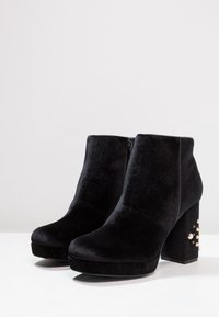 Botines de terciopelo negro con un tacón de plataforma chunky. El tacón presenta acentos y tachuelas en tono dorado. Textura suave en toda la bota.