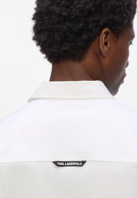 Weißes Hemd mit Kragen, ausgestattet mit einem schwarzen Etikett mit "KARL LAGERFELD" auf der Rückseite. Glattes Material und strukturiertes Design.