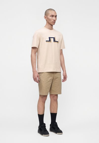 Άνδρας στέκεται φορώντας μπεζ γραφικό t-shirt, χακί σορτς, μαύρες κάλτσες και μαύρα αθλητικά παπούτσια σε απλό λευκό φόντο.