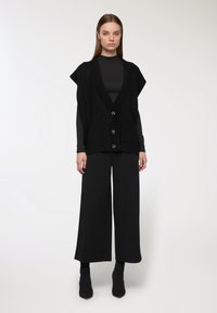 Czarna dzianinowa kamizelka z dekoltem w serek, krótkimi rękawami i czterema guzikami. Połączona z szerokimi czarnymi spodniami i dopasowanymi czarnymi botkami na obcasie.
