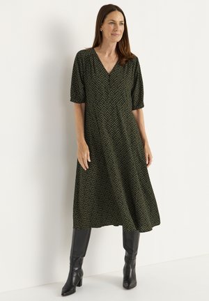 Robe verte à motifs avec des manches bouffantes courtes et un décolleté en V. Le tissu présente un design géométrique, et le mannequin porte des bottes noires jusqu'aux genoux.