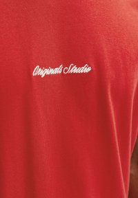 Punane puuvillane T-särk, mille seljal on valge käekirjas tikand tekstiga "Originals Studio", sile tekstuur ja lihtne disain.