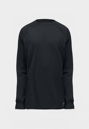 Jeune homme portant une chemise noire à manches longues, un pantalon ample vert olive et des baskets beige, regardant vers le bas les mains dans les poches.