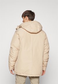 Beige polstret jakke med høy krage og hette, med en glatt, skinnende tekstur og elastiske mansjetter. Justerbar snor i nederkant.