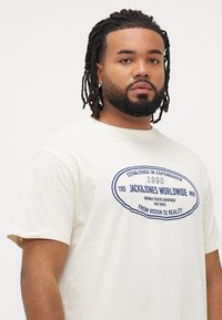 Mees, kellel on dredsid ja habe, kannab heledat valget T-särki, millel on ees "Jack & Jones Worldwide" sinise tekstiga logo, seisab otsa vaates.
