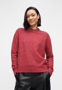 Pull rouge à col montant, fabriqué en tissu doux. Il présente des manches longues et des poignets côtelés, porté avec un pantalon en cuir noir.