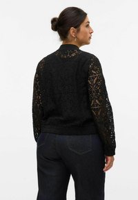 Veste en dentelle noire à manches longues, ornée de motifs floraux complexes et d'une taille ajustée. Associée à un jean en denim foncé.