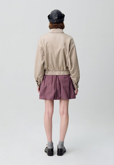 Giacca cropped beige con cintura elastica, abbinata a una gonna a scacchi rossa e bianca. Scarpe nere e calze grigie completano l'outfit.