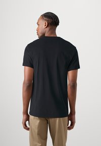 Schwarzes Baumwoll-T-Shirt mit kurzen Ärmeln, flachem Ausschnitt und lockerer Passform, von hinten gezeigt, kombiniert mit beigen Hosen.