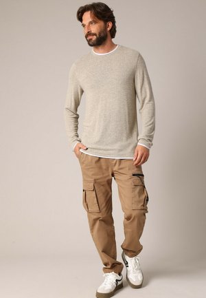 Chemise beige à manches longues avec un col blanc, associée à un pantalon cargo marron clair avec plusieurs poches, porté avec des baskets blanches et noires.