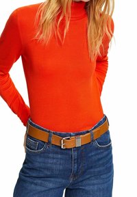 Pull à col roulé orange associé à un jean denim bleu. La ceinture en cuir marron est ornée d'une boucle argentée avec des rainures verticales.